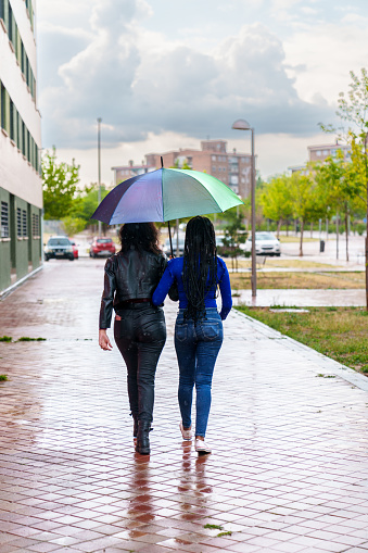 A vivid, heartwarming photo of two diverse friends, one Caucasian and the other African, linked arm in arm under a multicolored LGBT umbrella, joyfully walking through a rainy city
