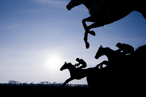 Silhouette of race horses jumping a fence against a setting sun, profile view with clear blue sky.
