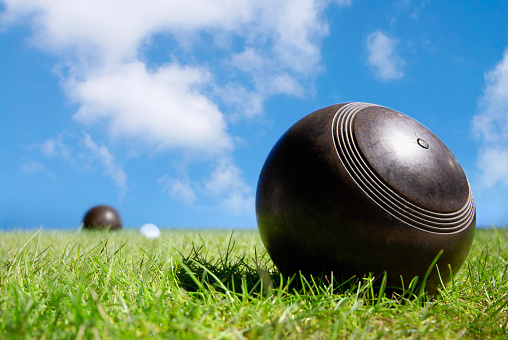 A game of lawn bowls being played on green grass. The balls are traditional, brown and made of wood. There is a blue sky above the balls and they rest around a white jack on the grass. The sun is bright and causes a harsh shadow from the ball in focus onto the grass.