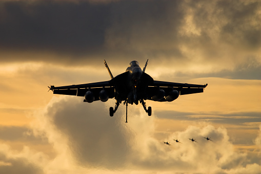 A naval jet fighter on approach for landing as more jets approach overhead.