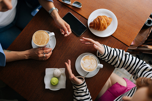 Closeup high-angle view of two unrecognizable happy female girlfriends sitting at table with coffee, croissant and macaroon in cafe, communicate and sharing stories spending relax in coffee shop.