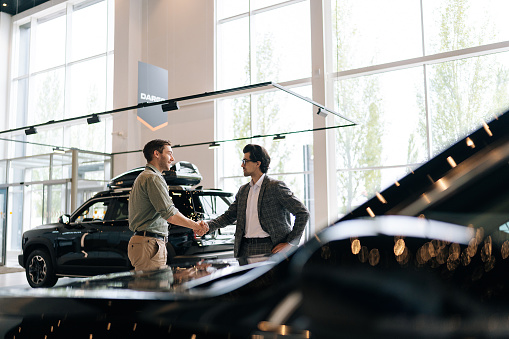 Remote view of young customer male thanking stylish dealer male wearing suit and eyeglasses for helping to choose car to buy in dealership center. Concept of choosing and buying new car at showroom
