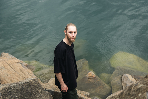 A young man with a beard and a bun in a gray T-shirt stands among the stones near the lake or the sea.