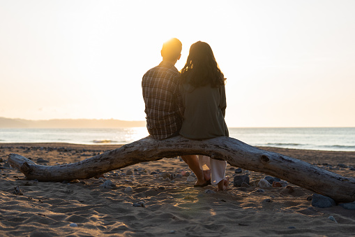 Silhouettes of a couple on beach at sunset , summer travel holidays. Romantic couple enjoying watching sunset sitting on tree trunk at the beach by the ocean.