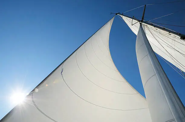 Sails of a yacht  billowing in the wind, against blue Mediterranean sky. Sun (and a lens flair) partially behind the sail.