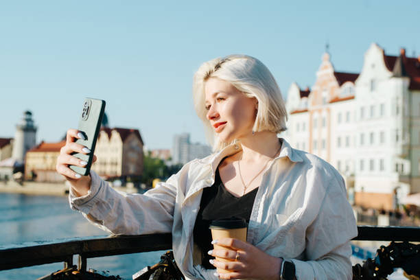 Pretty young woman taking selfie on smartphone, cute blonde teenager with cup of coffee using phone on bridge on sunny day stock photo