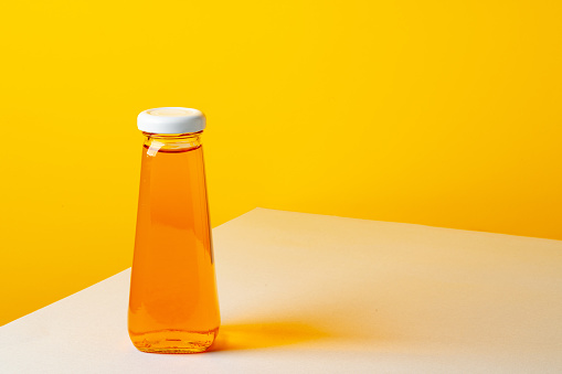 Glass bottle with juice on white table against yellow background close up