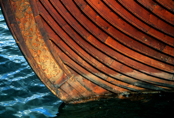A male viking warrior in traditional leather battle clothing and boots holding an axe and shouting, close to a moored longboat with lowered sail on a deserted rocky beach. With stormy sky and bright sun at dawn or dusk.