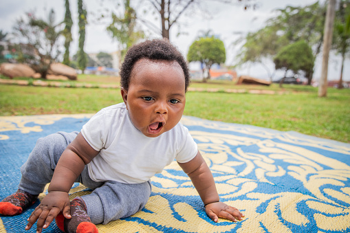 An African newborn baby yawns while sitting on a mat in a public park