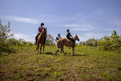 Two girls riding horses together outdoors on a meadow, their riding instructor is with them.