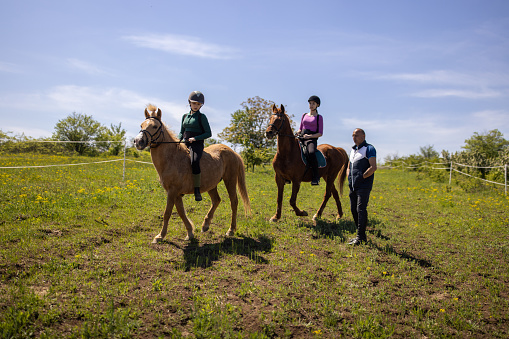 Two girls riding horses together outdoors on a meadow, their riding instructor is with them.