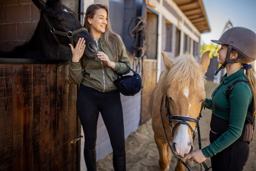 Woman and girl together with horses outdoors by the stable.