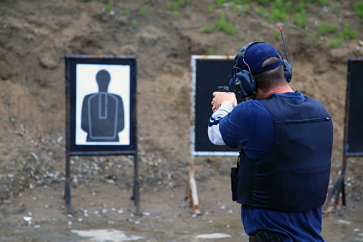 Picture of a man shooting shotgun in a practice field.  The man is wearing a kevlar vest and has earmuffs to protect him from the sound of the handgun. The shooting target is blur and visible in the background. 