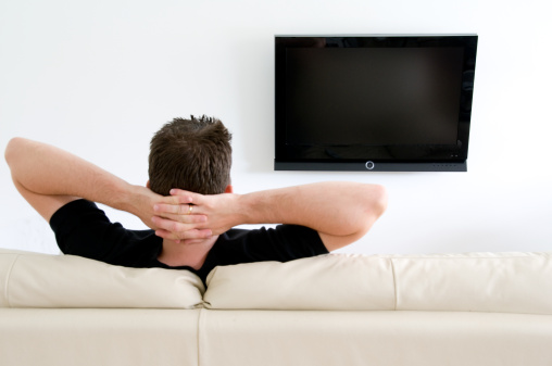 Man sitting on a sofa watching tv. Hands folded behind his neck - relaxing. Black screen.