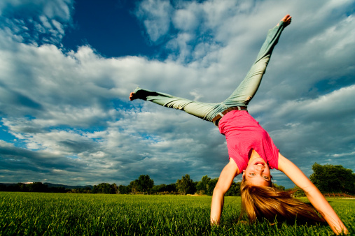 Pretty athletic young girl smiling and doing a cartwheel on a vast green lawn with a dramatic cloudscape in the background.  There are trees at the end of the field and the girl is wearing blue jeans and a casual pink top and is barefoot.