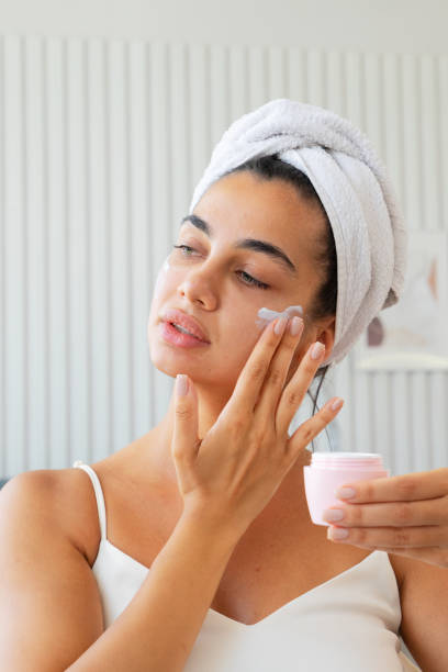 istock Charming brunette woman with towel wrapped on her head applying moisturizing face cream 1574310985