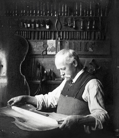 Vintage black and white image of a skilled luthier making a cello in his workshop. Probably in Mirecourt France. Circa 1900. Some dust and scratches which convey age of original image. 