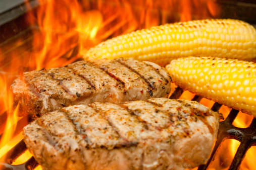 Thick juicy pork chops with grill marks and roasted corn on the cob on an old fashioned charcoal barbecue grill with big beautiful orange flames engulfing the food, searing in the juices and adding smokey flavor.