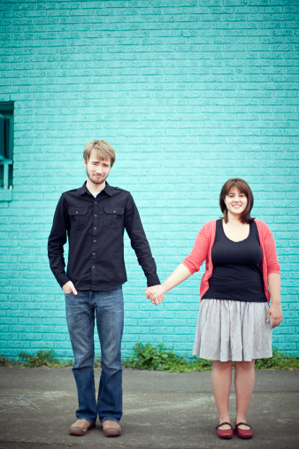 A young man and woman stand in an urban setting in front of a bright blue/turquoise wall holding hands and smiling.  Vertical with copy space.