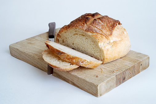 a round artisan loaf with two slices sitting on a wooden cutting board with a bread knife.  White background