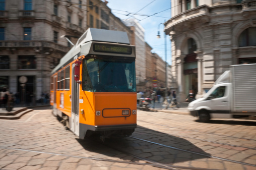 Motion blurred image of distinctive Milanese orange electric tram whizzing on its rails through the cobbled Piazza Cordusio in the beautiful capital city of Lombardy, Italy. ProPhoto RGB profile for maximumc color fidelity and gamut.