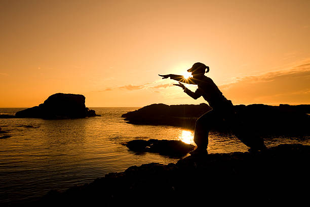Woman practicing meditation-martial arts on a rocky ocean shore with the warm colors of the Sun in the background.