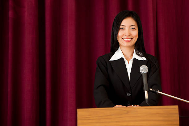 An attractive asian woman standing at a podium in front of a red stage curtain with a big smile. Politics, Education, or Business