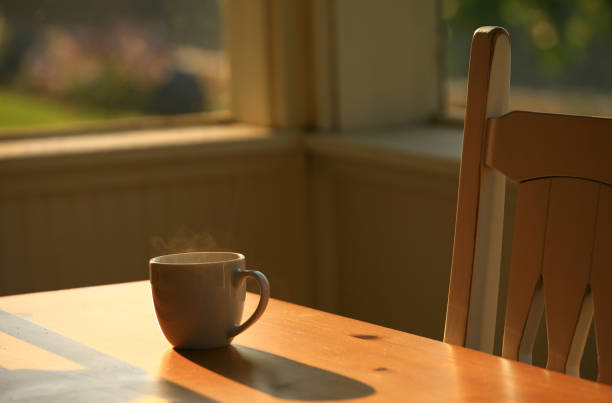 A hot cup of coffee in the veranda at a beach house. Coffee is backlit, hot, and steaming as it sits on a rustic wood table in a cabin. Themes include coffee, caffeine, cup, beverages, hot drinks, morning, relaxing, ceramic, porch, rustic, country, hot chocolate, espresso, Americano, latte, cappuccino, and steam. 