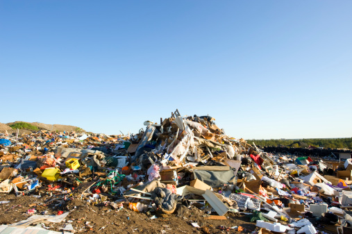A huge pile of garbage is in the middle of this photograph, spreading out in all directions.  Green grass can be seen behind the garbage on the right side of the image.  The sky above the landfill is a bluish-white near the horizon, fading to a darker blue near the top of the image.  Good copy space at top.