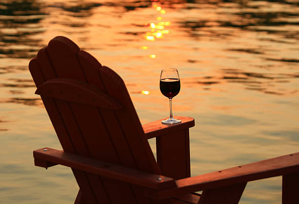 Adirondack Chair and Wine at Sunset By Lake A glass of wine on an adirondack chair by the lake. Thompson Okanagan, British Columbia. This relaxing and luxurious scene is in the South Okanagan region of British Columbia. However, it could be a relaxing scene on any summer lake. Additional themes include red wine, relaxation, holiday, summer, opulence, calm, serene, tranquility, quiet, nobody, peaceful, and solitude.  cottage life stock pictures, royalty-free photos & images