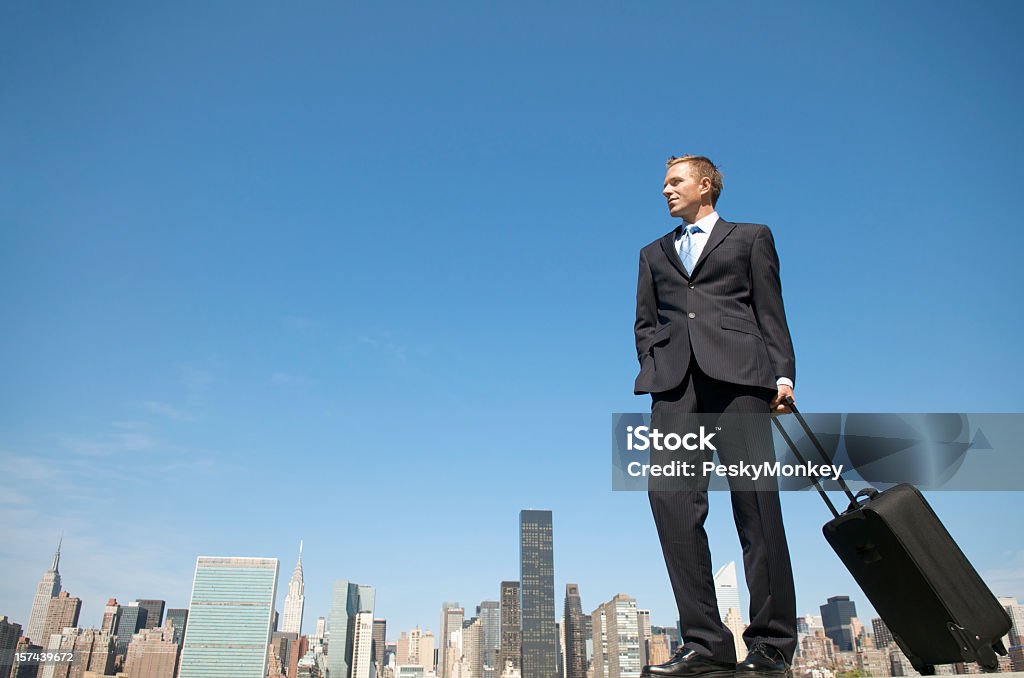 Young Businessman Standing with Suitcase above City Skyline Young businessman towering over city skyline holding his rolling suitcase Adult Stock Photo