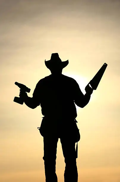 A silhouette of a construction worker holding tools. "Handyman" holding drill and hand-saw. Rear view. Vertical image. Caucasian cowboy tradesman is jack of all trades and also has tool belt with hammer. Image is designed to show a "gun fight," or western gun slinger theme. Except, of course, the gun fight is with tools. Themes include building, working, blue collar, renovation, construction, worker, evening, cowboy, wild west, carpenter, carpentry, trade, and building. 
