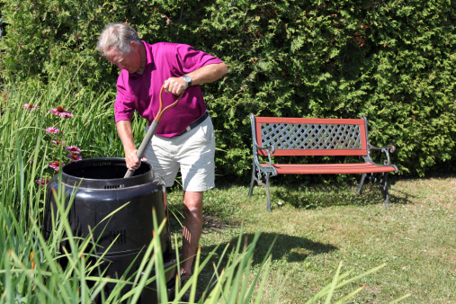 A DSLR photo of senior man holding a metal and wood shovel full of fresh compost over a black compost bin. There is flowers in background, it's a sunny summer day in the garden.