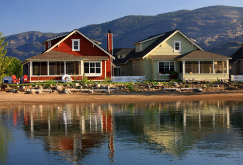Two colourful beach houses on a pristine lake in the Thompson Okanagan region of British Columbia, Canada. Lake houses or cottages feature Adirondack chairs and beautiful colours that are reflected in the lake. Houses also have screened porches or verandas and are in the 50s style of homes. 