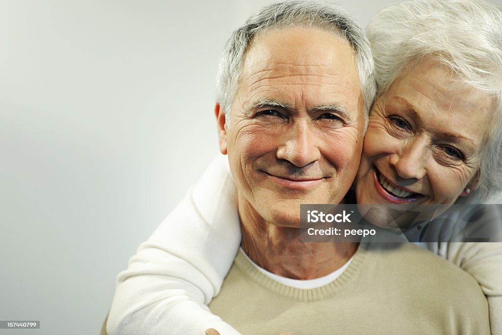 Senior Couple A close up head and shoulders portrait of a happy hetrosexual senior couple. The mature woman is wrapping her arms around the mature man in an affectionate loving embrace from behind and the couple are smiling cheerfully and looking at the camera. Both seniors are dressed in white and beige casual clothes.  Human Face Stock Photo