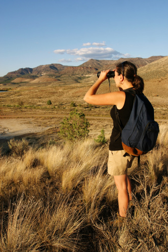 Woman hiking through desert wilderness with backpack, standing in tall grass, looking with binoculars