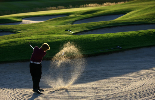 A young junior golfer blasts a ball out of a bunker on a beautiful golf course. Sand is backlit to provide drama. Themes include golf, golfer, boys, junior level athlete, golf course, action, motion, sports, leisure, golf resort, summer, swinging, athletic, teenager, back view, sand, airborne, and skill. 