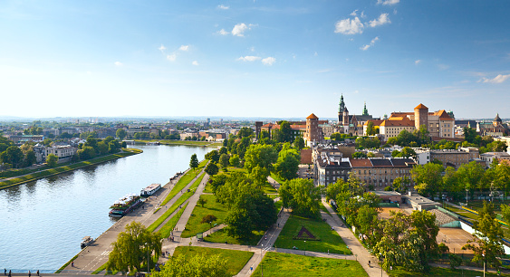 Daytime panorama of Krakow, Poland, featuring Wawel Castle.  The Vistula River flows along the left side of the image, and the castle is visible on the right, near the horizon.  There is a blue sky with sparse, wispy clouds above the horizon and a manicured lawn intersected by walking paths in the foreground.
