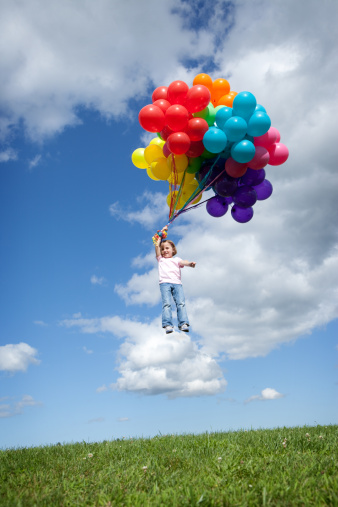 Color photo of a five-year-old girl smiling as she holds a bunch of helium balloons that are carrying her away into the blue sky.