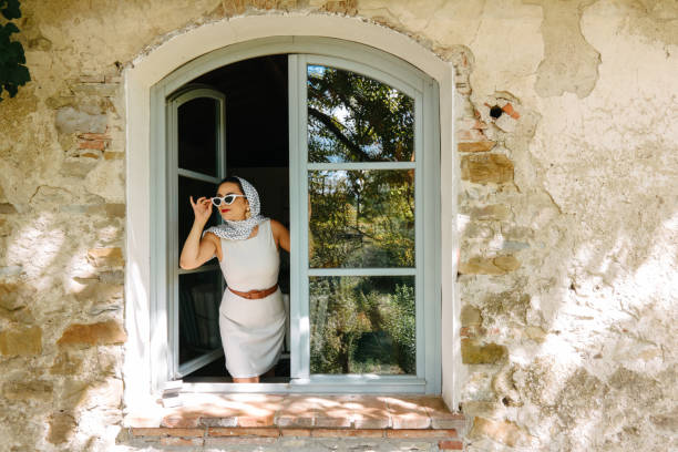 A young woman, well-dressed in retro clothes, looking out of the window of her old stone house in Anghiari in Italy. Traditional homes