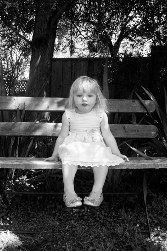 A little girl with pigtails in a summer white dress, offended, sits on a bench hugging her legs with her arms. Portrait of an offended child, sadness. Carefree childhood. toning.
