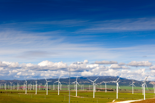 Outdoor photo of several wind turbines on a wind farm in southern Spain.  Each white turbine has three blades.  They are lined up in numerous rows.  The grass is mostly green with a few brown spots.  A few small trees stand near some of the turbines.  Green mountains with bare dirt spots are positioned in the background.  The sky has a gradual color change beginning with light blue at mountain level to bright blue at the top of the image.  There are cumulus clouds above the mountains in the image.