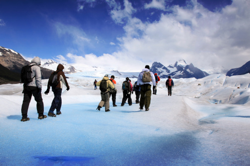 DSLR photo of a group of people hiking on the Perito Moreno glacier. The group is walking on blue ice and are wearing crampons not to slip. They are walking towards the mountain in the background of the picture. It is mostly a blue sky with few clouds. 