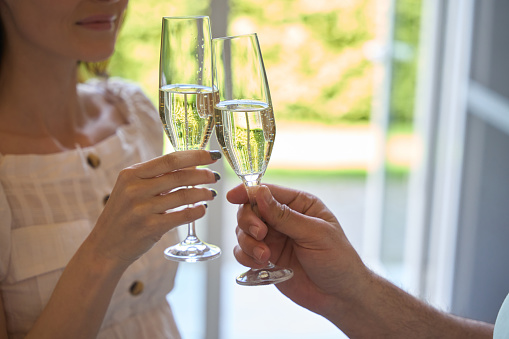 Cropped view of young woman with beautiful manicure clinking wineglasses with her man indoors