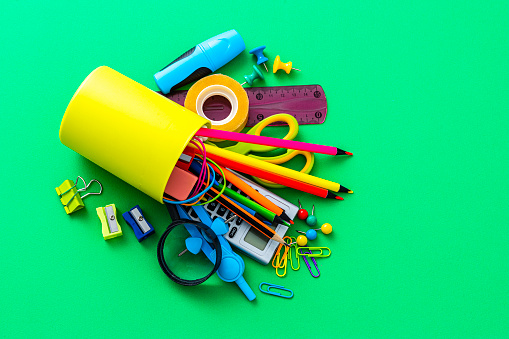 Overhead view of a desk organizer full of school supplies laying on green background. The composition includes a drawing compass, color pencils, paint brushes, paper clips, pencil sharpener, eraser, crayons, pocket calculator, thumbtack, rubber bands, notepad, stapler, adhesive tape, felt tip pen and scissors.
