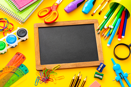 Composition of multi colored school supplies arranged all around a chalkboard shot on yellow background. The composition includes a drawing compass, color pencils, paint brushes, paper clips, pencil sharpener, eraser, crayons, pocket calculator, thumbtack, rubber bands, notepad, stapler, adhesive tape, felt tip pen and scissors.