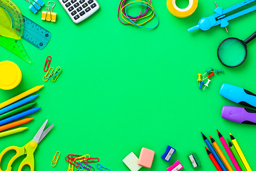 Composition of multi colored school supplies arranged all around the border of a green background making a frame and leaving copy space at the center. The composition includes a drawing compass, color pencils, paint brushes, paper clips, pencil sharpener, eraser, crayons, pocket calculator, thumbtack, rubber bands, notepad, stapler, adhesive tape, felt tip pen and scissors.
