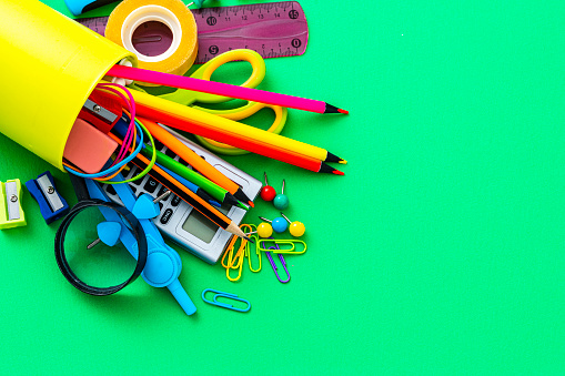 Overhead view of a desk organizer full of school supplies laying on green background. Copy space. The composition includes a drawing compass, color pencils, paint brushes, paper clips, pencil sharpener, eraser, crayons, pocket calculator, thumbtack, rubber bands, notepad, stapler, adhesive tape, felt tip pen and scissors.