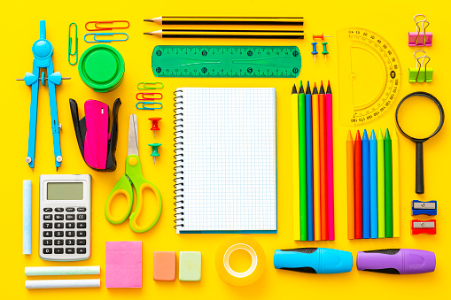 Composition of multi colored school supplies shot on yellow background. The composition includes a drawing compass, color pencils, paint brushes, paper clips, pencil sharpener, eraser, crayons, pocket calculator, thumbtack, rubber bands, notepad, stapler, adhesive tape, felt tip pen and scissors.