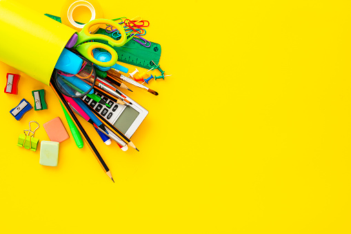 Overhead view of a desk organizer full of school supplies laying on yellow background. Copy space. The composition includes a drawing compass, color pencils, paint brushes, paper clips, pencil sharpener, eraser, crayons, pocket calculator, thumbtack, rubber bands, notepad, stapler, adhesive tape, felt tip pen and scissors.